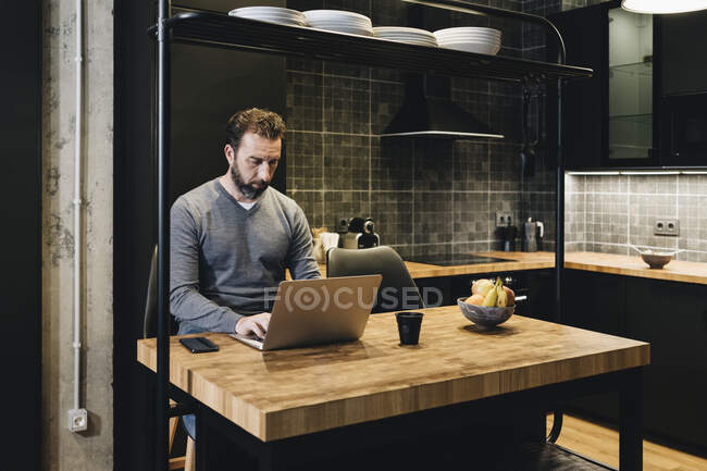 Mature businessman working in kitchen, using laptop — Stock Photo