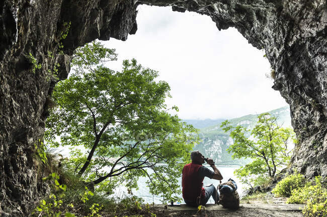Männlicher Wanderer trinkt, während er mit Rucksack am Höhleneingang sitzt — Stockfoto