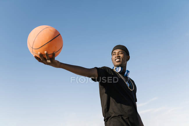 Молодий чоловік тримає баскетбол, стоячи на тлі чистого блакитного неба під час сонячного дня — стокове фото