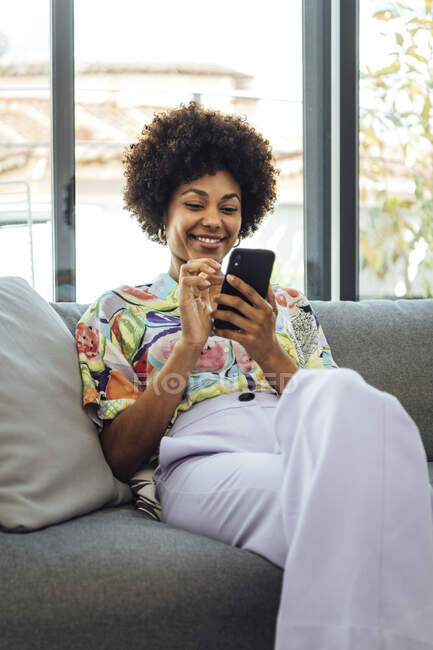 Feliz media mujer adulta usando el teléfono inteligente mientras está sentado en el sofá en el ático moderno - foto de stock
