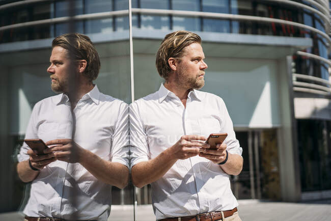 Geschäftsmann in der Stadt lehnt an Glasfront und hält Smartphone — Stockfoto