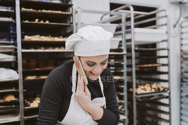 Happy female baker using smart phone in kitchen at bakery — Stock Photo