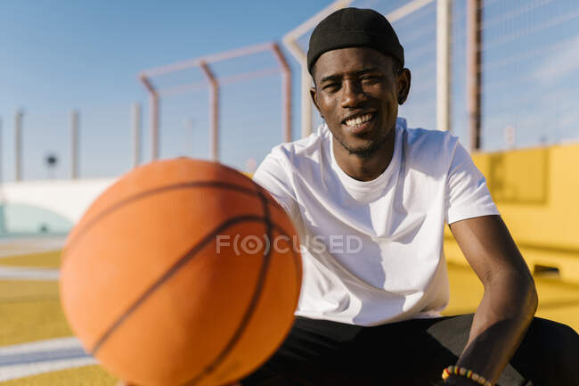 Smiling young man holding basketball while sitting in court during sunny day — Stock Photo