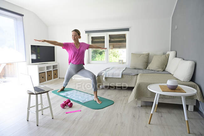 Mujer madura con laptop practicando yoga en colchoneta de gimnasio en salón - foto de stock
