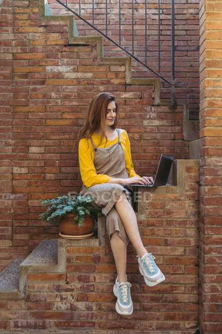 Giovane donna con i capelli lunghi utilizzando il computer portatile mentre seduto su passi contro muro di mattoni — Foto stock