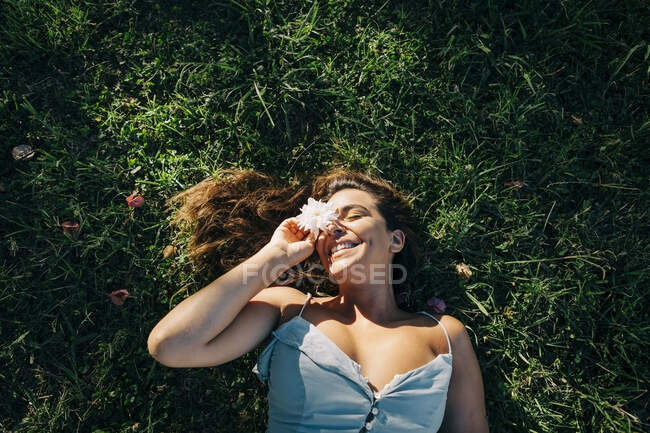 Mulher sorridente com os olhos fechados segurando flor enquanto relaxa em terra gramada no parque — Fotografia de Stock