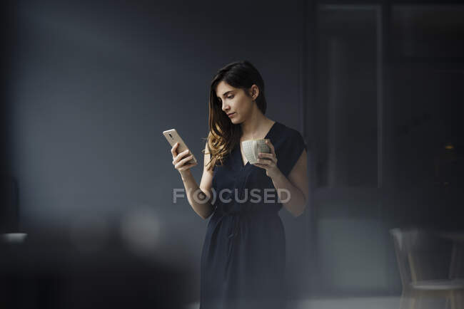 Портрет серйозної молодої жінки з чашкою чаю у високій кімнаті, яка дивиться на мобільний телефон. — стокове фото