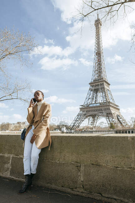 Mujer usando un teléfono inteligente mientras está de pie en el puente con la Torre Eiffel en segundo plano contra el cielo, París, Francia - foto de stock
