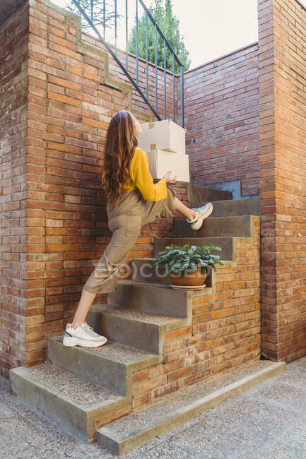 Giocoso giovane donna che trasporta scatole mentre si muove su gradini da muro di mattoni — Foto stock