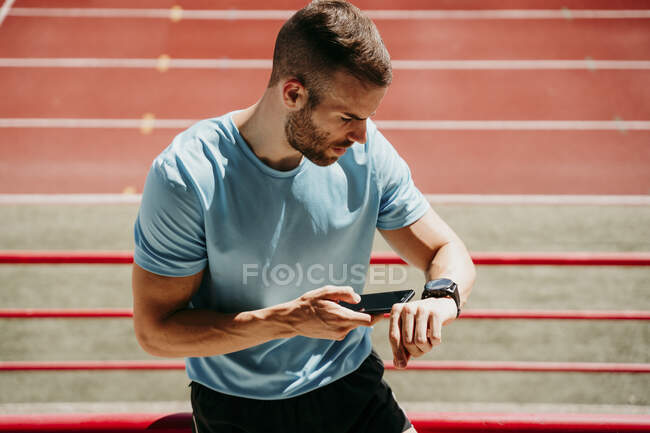 Atleta masculino en el estadio comprobando smartwatch y smartphone - foto de stock