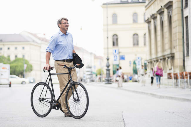 Empresário sorrindo olhando para longe enquanto caminhava com bicicleta na estrada na cidade — Fotografia de Stock