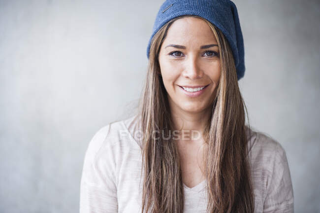Mujer feliz con el pelo largo marrón usando sombrero de punto contra la pared gris - foto de stock
