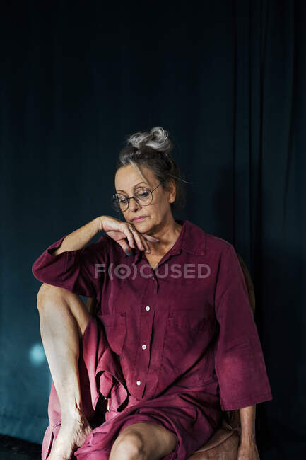 Mujer mayor pensativa con anteojos sentados en la silla contra la cortina en casa - foto de stock