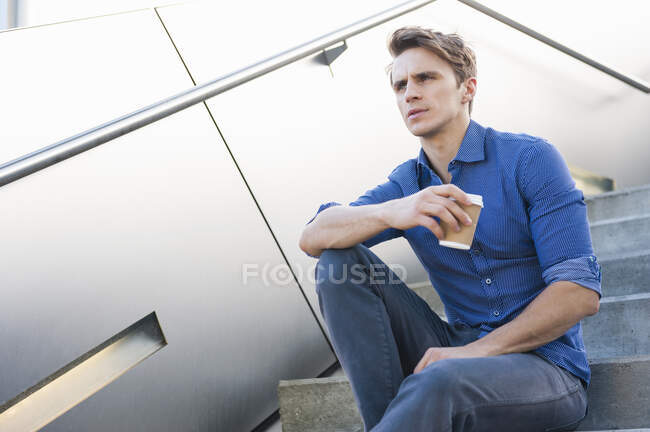 Pensativo hombre de negocios sosteniendo café mientras está sentado en los escalones - foto de stock