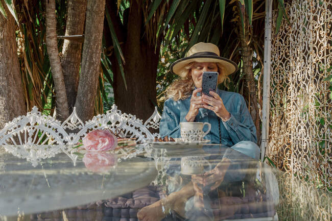 Seniorin sitzt auf Gartenbank und entspannt sich mit Tasse Tee und Keksen per Smartphone — Stockfoto