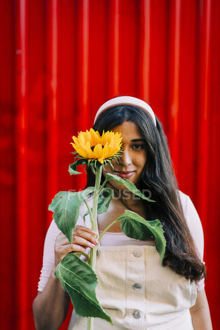 Young woman in front of red wall, covering eye with sunflower — Stock Photo