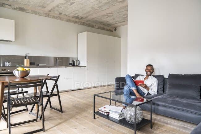 Uomo ridente con libro seduto sul divano in appartamento moderno — Foto stock