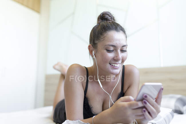 Sorridente giovane donna godendo la musica mentre si utilizza smart phone a letto a casa — Foto stock
