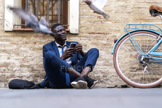 Joven hombre de negocios con smartphone sentado en el suelo de la ciudad - foto de stock