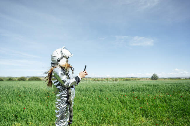Vista lateral de la chica en traje espacial utilizando el teléfono inteligente mientras está de pie en la hierba - foto de stock