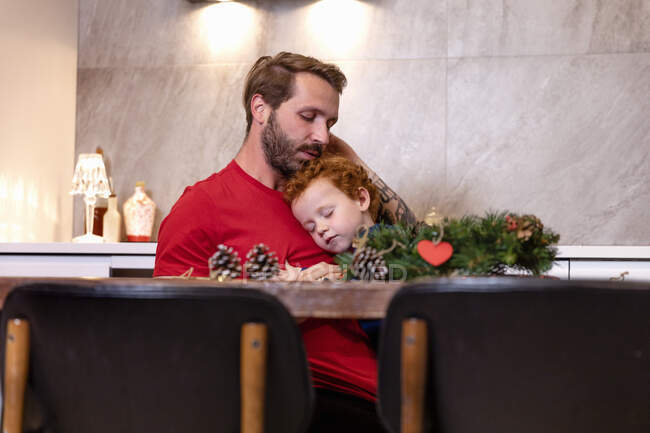 Father and son with Christmas wreath on table sleeping in kitchen at home — Stock Photo