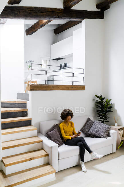 Jeune femme assise sur le canapé à la maison en utilisant un téléphone mobile — Photo de stock