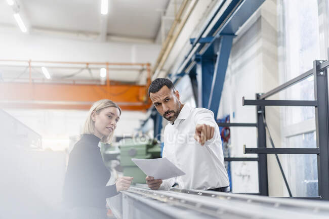 Geschäftsmann und Frau mit Papieren untersuchen Metallstäbe in Fabrikhalle — Stockfoto