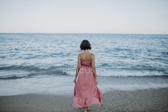Женщина в платье, идущая к морю на пляже против ясного неба во время заката — стоковое фото