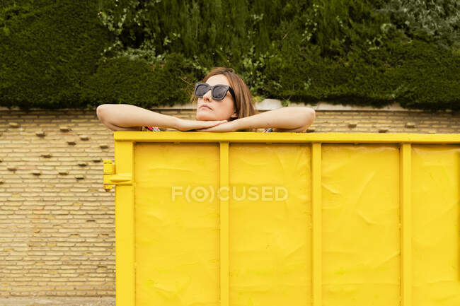 Woman wearing sunglasses, leaning on edge on yellow container — Stock Photo