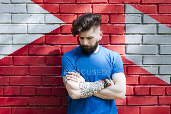Joven hombre con tatuaje de pie delante de la pared de ladrillo rojo con los brazos cruzados - foto de stock