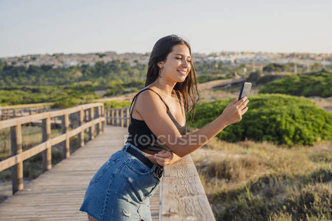 Ragazza adolescente prendendo selfie attraverso smart phone mentre in piedi sul lungomare in spiaggia — Foto stock
