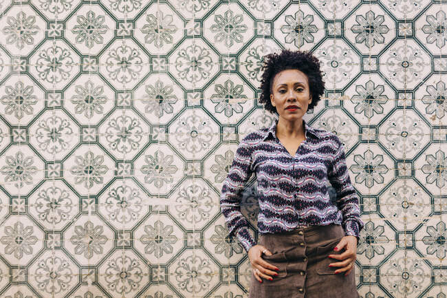 Elegante mujer adulta mediana con el pelo rizado de pie contra la pared de azulejos - foto de stock