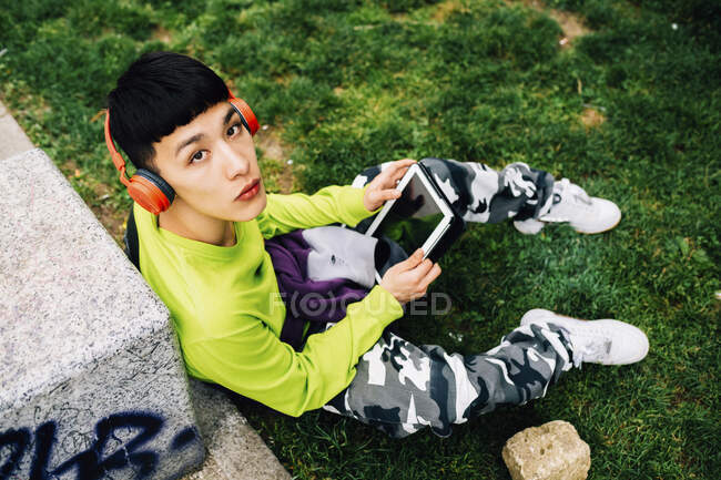 Homem jovem usando fones de ouvido usando tablet digital enquanto sentado em terra gramada — Fotografia de Stock