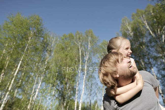 Feliz padre apoyando a su hija contra los árboles en el parque durante el día soleado - foto de stock