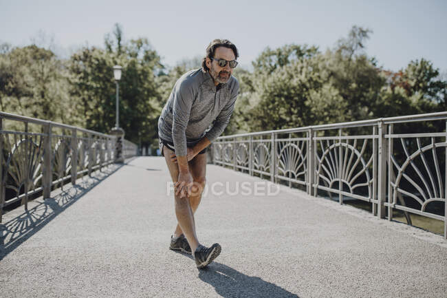 Homem usando óculos de sol esticando a perna enquanto estava em pé na passarela durante o dia ensolarado — Fotografia de Stock