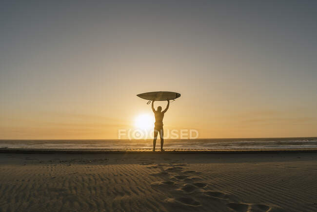 Surfista regge la tavola da surf sulla spiaggia, guardando il tramonto — Foto stock