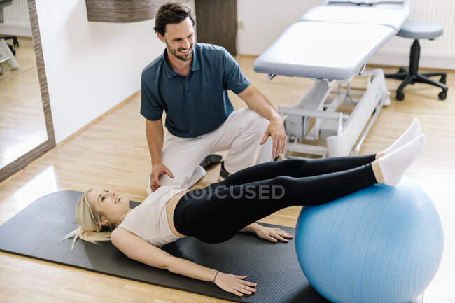 Physiotherapist assisting female patient, practicing with fitness ball, lying on gym mat — Stock Photo