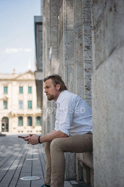 Homme d'affaires assis dans la niche d'un bâtiment tenant smartphone — Photo de stock
