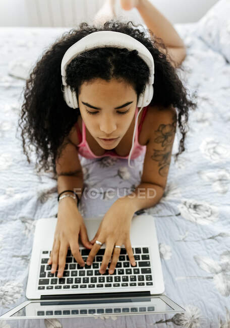 Beautiful young woman lying in bed at home wearing headphones and using laptop — Stock Photo