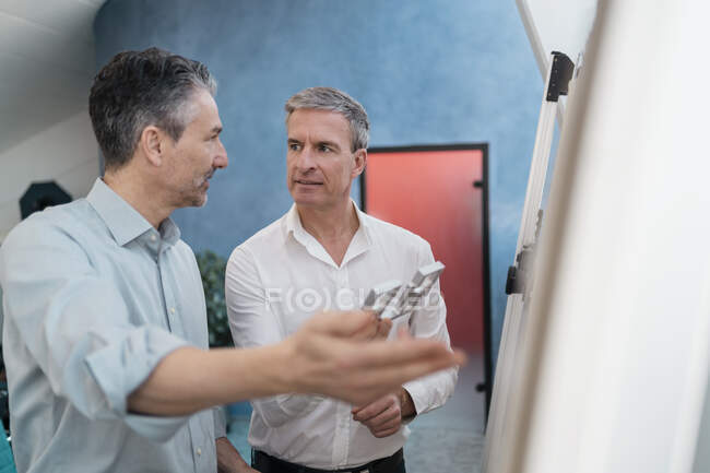 Mature male coworkers discussing over equipment while standing by whiteboard during meeting in office — Stock Photo