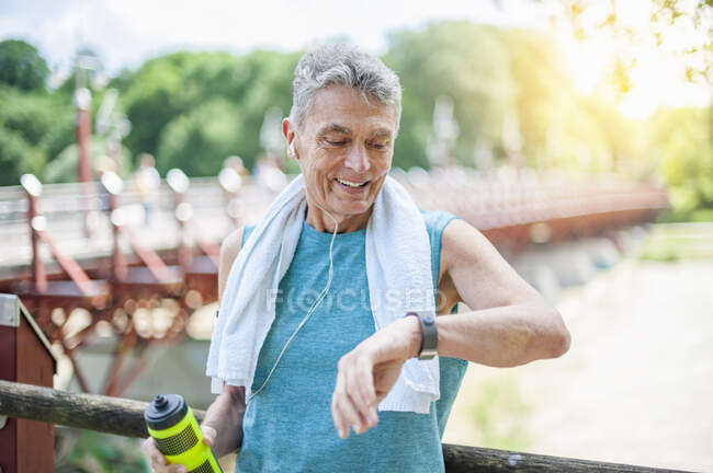 Улыбающийся активный пожилой человек смотрит на умные часы, стоя в парке — стоковое фото