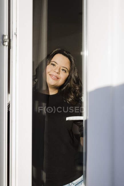 Задумчивая женщина с синдромом Дауна улыбается, глядя в окно дома — стоковое фото