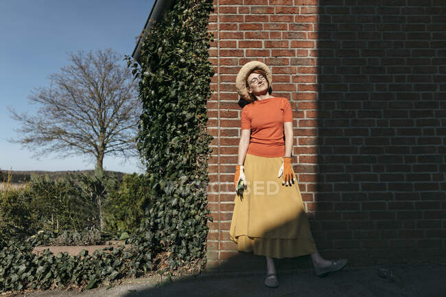 Портрет зрілої жінки з садівничими рукавицями та обрізкою, що стоїть перед цегляною стіною і насолоджується сонячним світлом. — стокове фото
