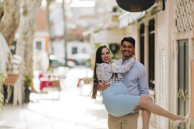 Hombre feliz llevando a la mujer embarazada en la ciudad durante el día soleado - foto de stock