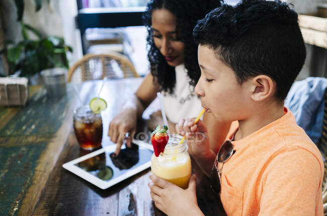 Vue latérale du garçon jouissant smoothie par mère assis avec tablette numérique au restaurant — Photo de stock