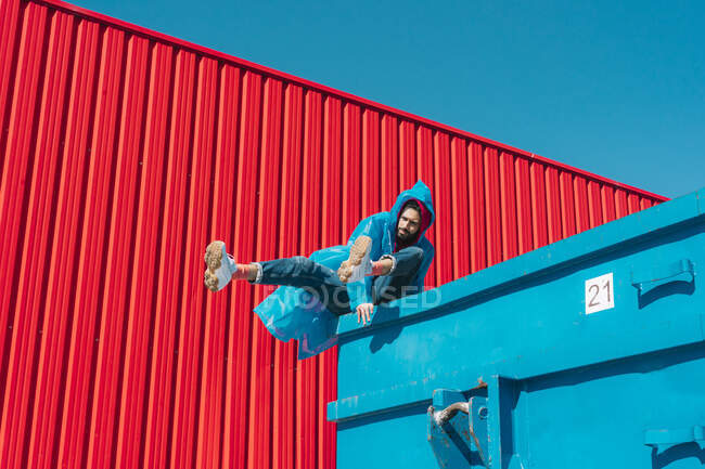 Joven vistiendo impermeable balanceándose en el borde del contenedor azul frente a la pared roja - foto de stock