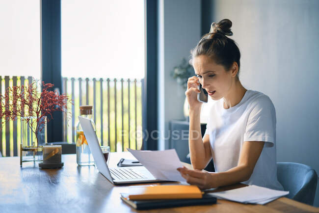 Деловая женщина держит документ разговаривая по смартфону на рабочем столе в домашнем офисе — стоковое фото