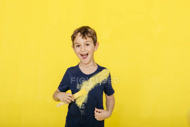 Portrait de garçon joyeux jouant avec le pinceau tout en se tenant debout sur fond jaune — Photo de stock