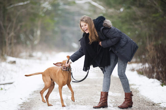 Cheerful young woman playing with dog on road in forest during winter — Stock Photo