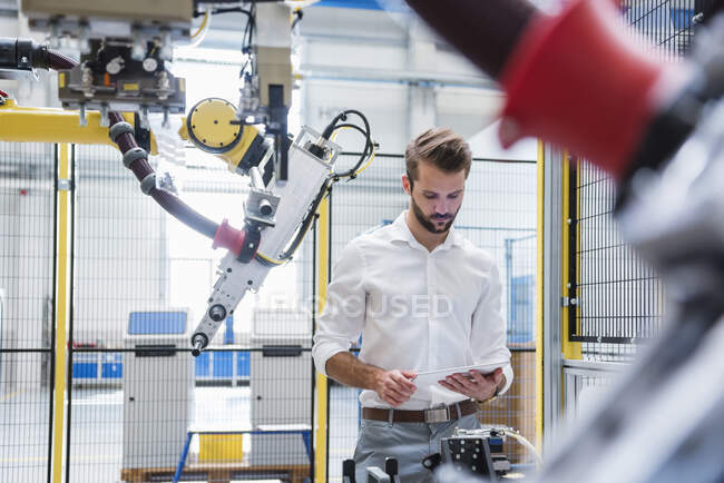 Молодой эксперт-мужчина, использующий цифровой планшет, стоя рядом с роботизированной рукой в промышленности — стоковое фото